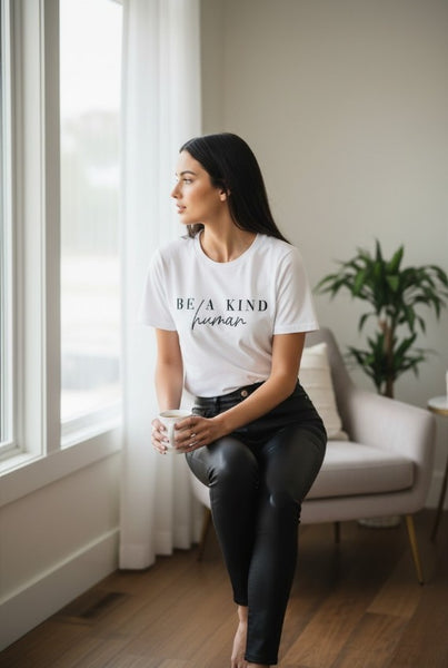 Woman sitting on a chair holding a mug, wearing a white t-shirt with text, in a room with a window and plant.