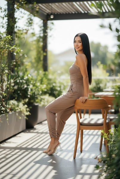 Collab Boutique taupe strapless ruched bodice jumpsuit with tapered legs and side pockets, worn by a model sitting on a chair outdoors.